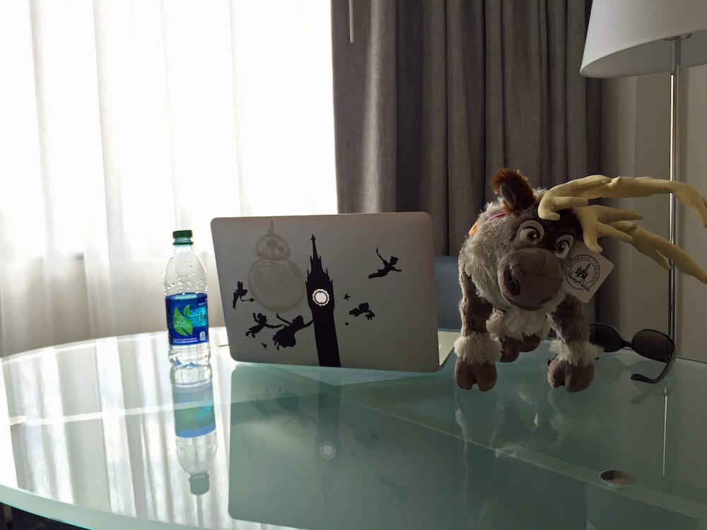 Glass desk top with a water bottle, Sven reindeer stuffed animal and macbook sitting on it.