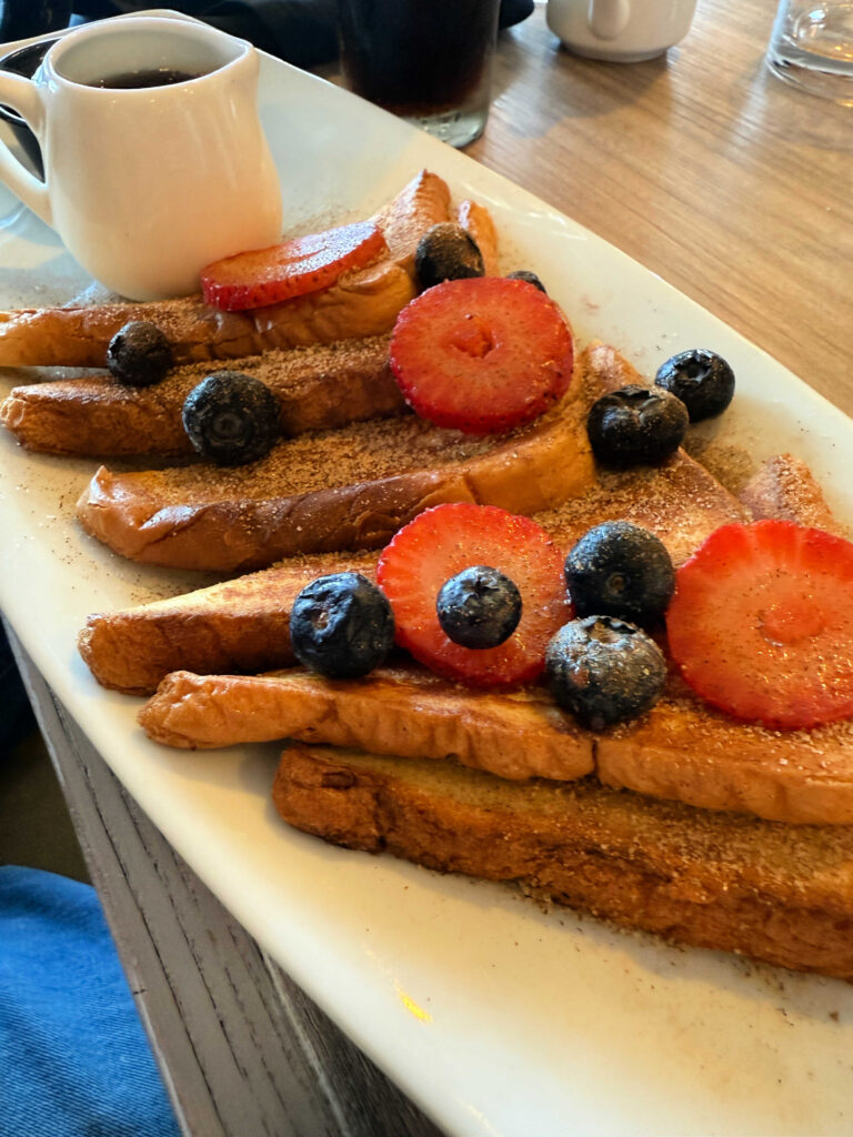 French toast covered in strawberries and blue berries on a white plate with a side of syrup